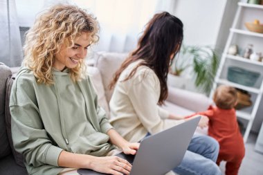 Bilgisayarlı sarışın kadına odaklan. Bulanık partnerinin yanındaki koltukta oturan bebeğine, ailesine.