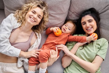 Çekici mutlu lgbt çifti kanepeye uzanmış ellerinde bebek mandalinalarıyla, aile kavramı