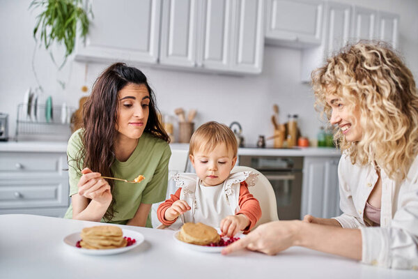 cheerful bonded lesbian couple having breakfast with their small pretty daughter, family concept