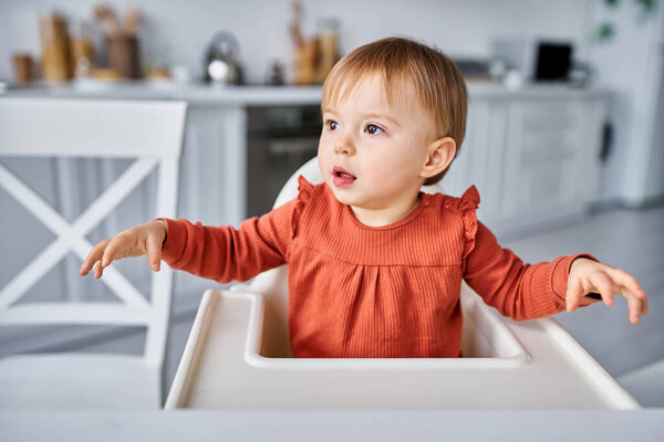 cute pretty toddler girl in orange sweater sitting on high chair at breakfast and looking away