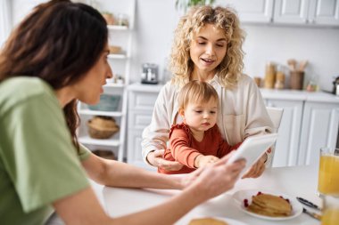 charming lgbt couple having breakfast with their baby girl and looking at tablet, family concept clipart