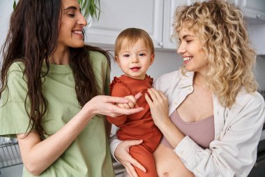 bonded cheerful lesbian couple in homewear posing happily with their baby girl, modern parenting clipart