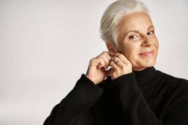 happy middle aged business woman in elegant attire wearing hoop earrings on grey background clipart