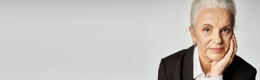 Güzel ve gri saçlı orta yaşlı kadın zarif giyinmiş gri arka planda poz veriyor.