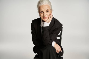 Güzel ve gri saçlı orta yaşlı kadın zarif giyinmiş gri arka planda poz veriyor.