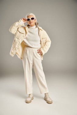 Güzel ve gri saçlı orta yaşlı kadın zarif giyinmiş gri arka planda poz veriyor.