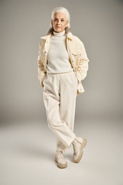 Güzel ve gri saçlı orta yaşlı kadın zarif giyinmiş gri arka planda poz veriyor.