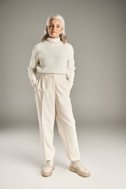 Güzel ve gri saçlı orta yaşlı kadın zarif giyinmiş gri arka planda poz veriyor.