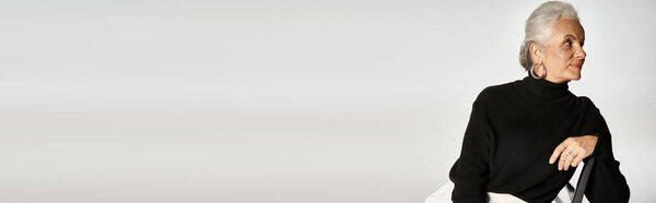 charming middle aged woman in smart casual attire and hoop earrings sitting on chair on grey, banner