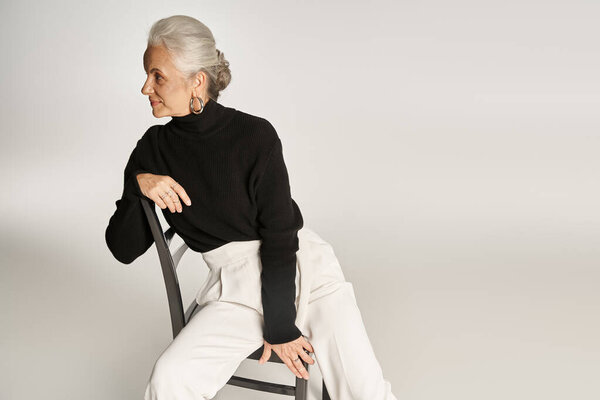 attractive middle aged woman in smart casual attire and hoop earrings sitting on chair on grey