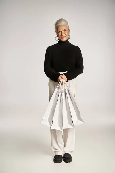 stock image cheerful middle aged woman in elegant attire and hoop earrings standing with shopping bags on grey