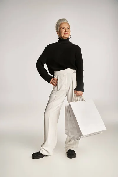 stock image satisfied middle aged woman in elegant attire and hoop earrings standing with shopping bags on grey