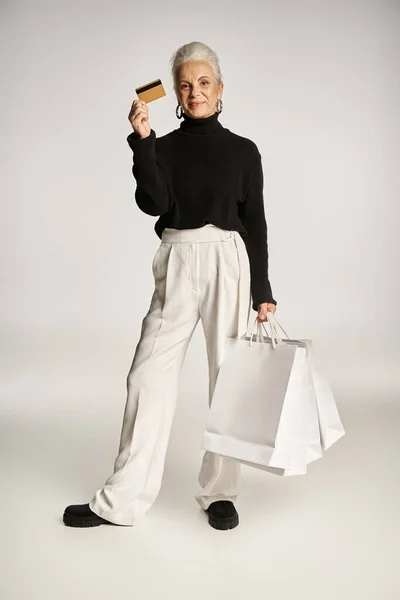 stock image satisfied middle aged woman in elegant attire holding credit card and shopping bags on grey backdrop