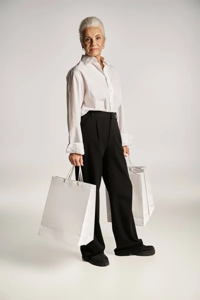 stock image full length, middle aged woman in white shirt and black pants standing with shopping bags on grey