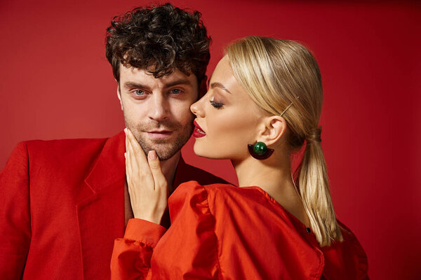 blonde woman with red lips touching face of handsome man in stylish attire on vibrant backdrop