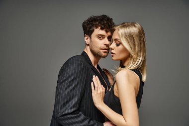 Siyah elbiseli sarışın kadın, gri arka planda güzel görünümlü bir adamı baştan çıkarıyor.