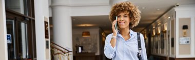Mutlu Afro-Amerikalı kadın otel lobisinde yürürken telefonla konuşuyor, yatay afiş.