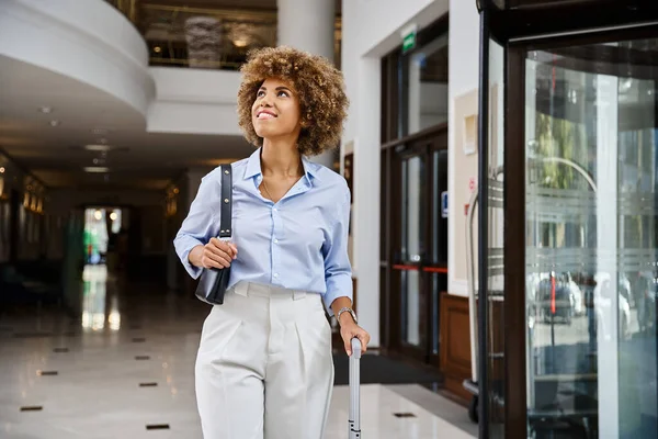 cheerful african american female traveler walking at hotel lobby entrance with her suitcase