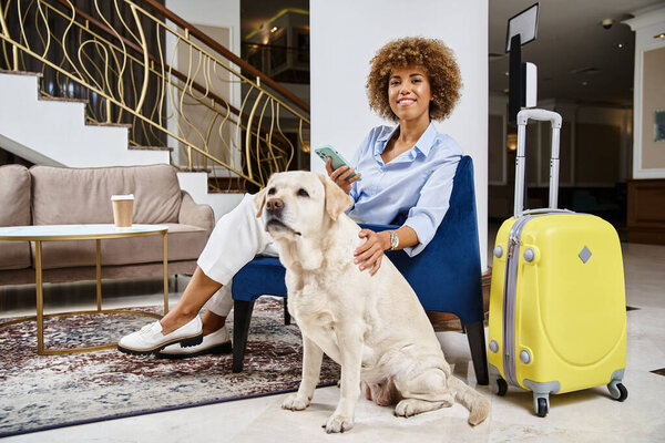 joyful african american woman with smartphone sitting near labrador in lobby of pet-friendly hotel