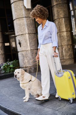 positive african american woman with dog and luggage standing near entrance of pet friendly hotel clipart