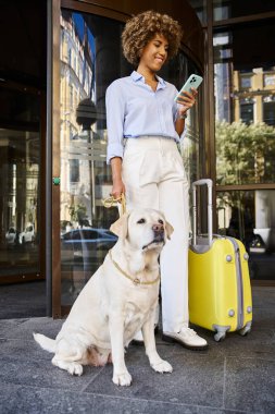 Gülümseyen Afrikalı Amerikalı bir gezgin hayvan dostu bir otel girişinde köpeğin yanında akıllı telefon kullanıyor.