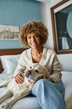 Genç ve neşeli Afro-Amerikalı bir kadın hayvan dostu bir otel odasında Labrador 'a sarılıyor.