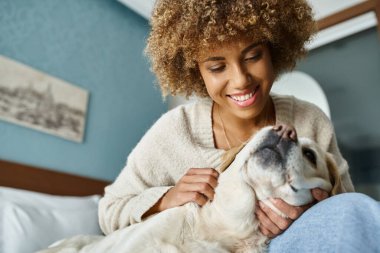 Evcil hayvan dostu bir otel odasında Labrador 'a sarılan neşeli, Afro-Amerikalı bir kadın.