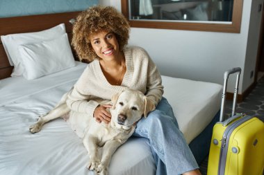 Mutlu Afro-Amerikalı kadın hayvan dostu otel odasında valizlerin yanında Labrador 'a sarılıyor.