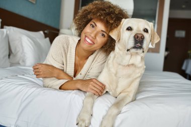 Gleeful african american woman with her labrador lying on a bed in a pet-friendly hotel, travel clipart