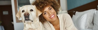 happy african american woman with her labrador lying on a bed in a pet-friendly hotel, banner clipart