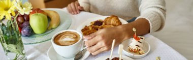 cropped banner of african american woman enjoying cappuccino and pastries in hotel room clipart