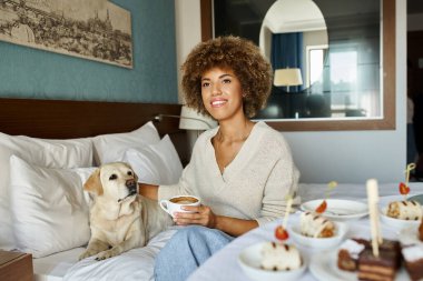 Neşeli Afrikalı Amerikalı kadın kapuçino tutuyor ve labrador köpeğini evcil hayvan dostu otelinde kucaklıyor.