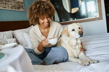 Neşeli Afrikalı Amerikalı kadın elinde fincanla labrador köpeğini evcil hayvan dostu otel odasında kucaklıyor.