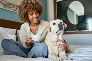 Mutlu Afro-Amerikan kadın elinde bir fincan kahve tutarken ve labradorunu evcil hayvan dostu otelinde kucaklarken.