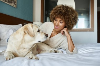 Neşeli Afrikalı Amerikalı kadın yatakta uzanıyor ve evcil hayvan dostu otelinde labrador köpeğine sarılıyor.