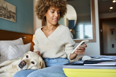Mutlu Afro-Amerikalı kadın, labrador köpeğiyle birlikte hayvan dostu bir otelde açık bagajın yanında oturuyor.