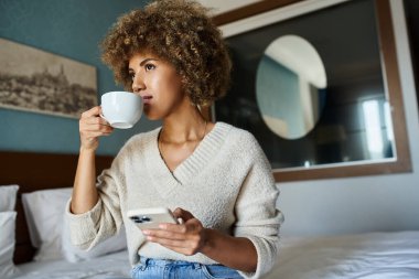 Kıvırcık saçlı Afrikalı Amerikalı kadın otel odasında akıllı telefon kullanırken kahvesini yudumluyor, seyahat ediyor.