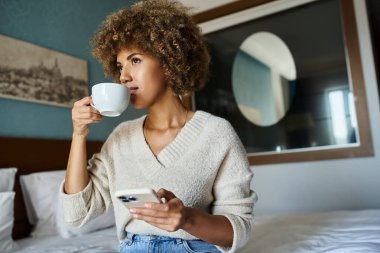 curly haired african american woman sipping coffee while using smartphone in hotel room, travel clipart