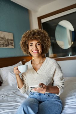 Kıvırcık saçlı ve mutlu Afro-Amerikalı kadın otel odasında elinde akıllı telefonla kahve yudumluyor.