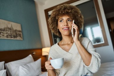 Mutlu Afro-Amerikalı kadın otel odasında telefonla konuşurken elinde bir fincan kahve tutuyor.