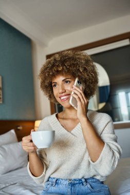 Neşeli Afro-Amerikalı kadın otel odasında telefonla konuşurken elinde bir fincan kahve tutuyor.