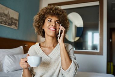 Pozitif Afro-Amerikalı kadın otel odasında telefonla konuşurken elinde bir fincan kahve tutuyor.