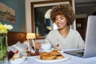 young african american woman using her laptop near breakfast in hotel, room service and convenience clipart