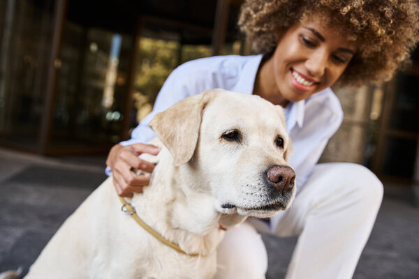 Smiling and curly african american woman cuddling labrador outside a pet-friendly hotel
