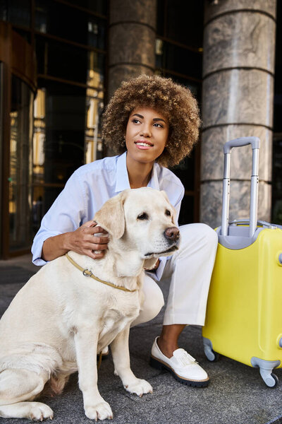 Smiling african american woman cuddling labrador near luggage outside a pet-friendly hotel
