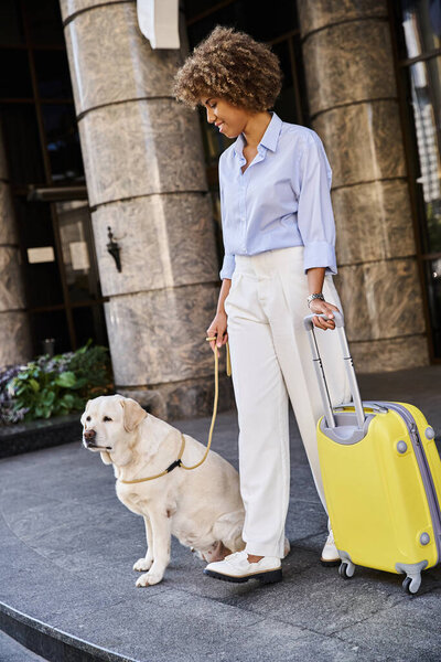 positive african american woman with dog and luggage standing near entrance of pet friendly hotel