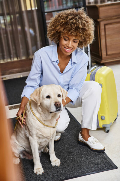 cheerful african american woman cuddling labrador near suitcase in lobby of pet-friendly hotel