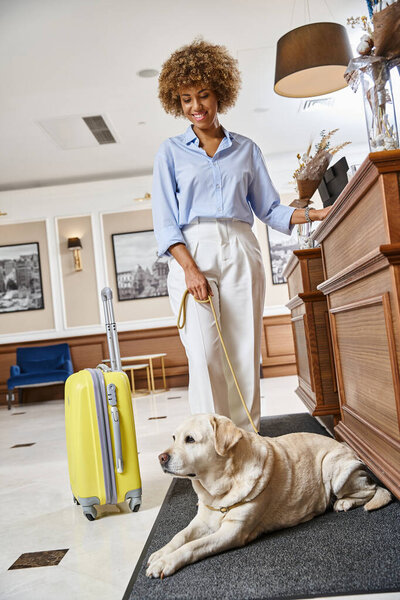 cheerful african american woman with her labrador checking in at a pet-friendly hotel on reception