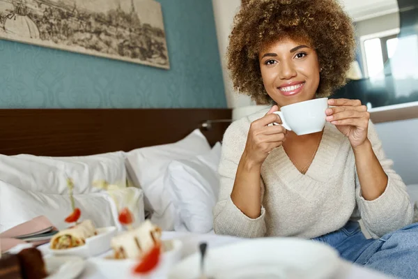 Stock image joyful african american woman holding cup and sitting on bed in hotel room, room service and comfort