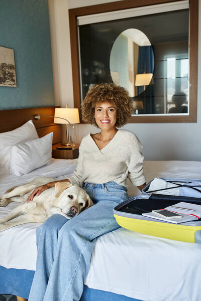 jolly african american woman sitting with labrador dog near open luggage in a pet-friendly hotel